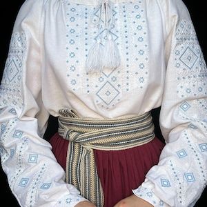 Puede incluir: Blusa blanca bordada con patrones geom&eacute;tricos azules y blancos. La blusa tiene una borla blanca en el escote y se lleva con un cintur&oacute;n a rayas marrones y blancas.