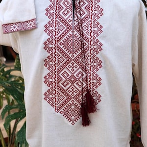 May include: An off-white long-sleeved shirt featuring a central vertical panel of intricate geometric embroidery in deep red thread. The shirt has a mandarin collar with a split neckline and two long red cords ending in matching tassels.