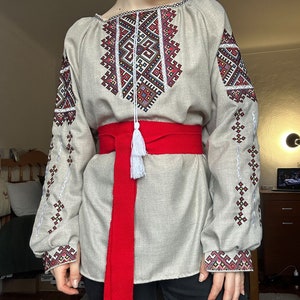 May include: A beige linen shirt with embroidered geometric patterns in red, black, and white. The shirt has long sleeves and a red sash with a white tassel.
