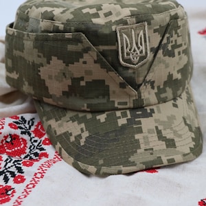 Pode incluir: Um boné militar de camuflagem com um emblema bordado azul e amarelo do brasão de armas ucraniano na frente. O boné está sobre um tecido branco bordado com flores vermelhas.