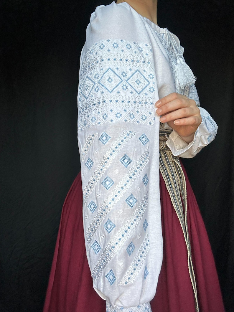 Puede incluir: Blusa blanca bordada con patrones geom&eacute;tricos azules y blancos. La blusa tiene mangas largas y un detalle de borla en el cuello.