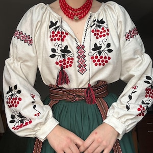 Pode incluir: Blusa bordada creme com desenhos florais vermelhos e pretos, mangas compridas e decote em V. A blusa é combinada com uma saia verde e um cinto castanho com detalhes vermelhos. Um colar de contas vermelho completa o look.
