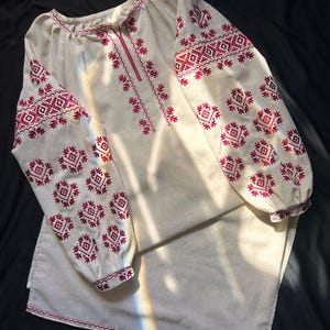 May include: A long-sleeved beige tunic featuring intricate red geometric embroidery patterns along the neckline, chest, and sleeves. The garment has a round neckline, gathered sleeve cuffs, and a straight hemline with matching red stitched detailing.