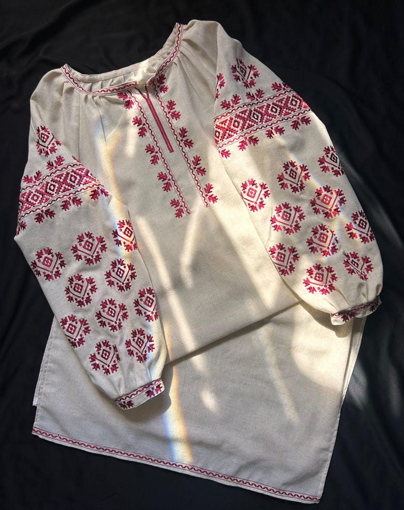 May include: A long-sleeved beige tunic featuring intricate red geometric embroidery patterns along the neckline, chest, and sleeves. The garment has a round neckline, gathered sleeve cuffs, and a straight hemline with matching red stitched detailing.