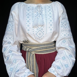 Puede incluir: Blusa blanca bordada con patrones geom&eacute;tricos azules y blancos, con un lazo de borla blanco en el cuello. La blusa se lleva con un cintur&oacute;n a rayas marr&oacute;n y blanco y una falda burdeos.