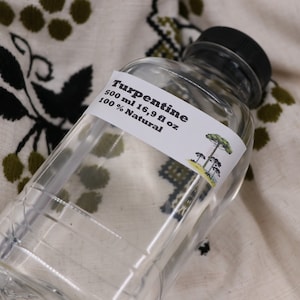 May include: A clear plastic bottle of turpentine with a black lid. The label reads "Turpentine 500 ml 16.9 fl oz 100% Natural" and features a drawing of pine trees.