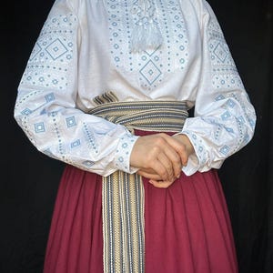Puede incluir: Una blusa blanca bordada con motivos geom&eacute;tricos azules y una borla blanca. La blusa se lleva con una falda color burdeos y un cintur&oacute;n tejido con rayas beige, azules y negras.