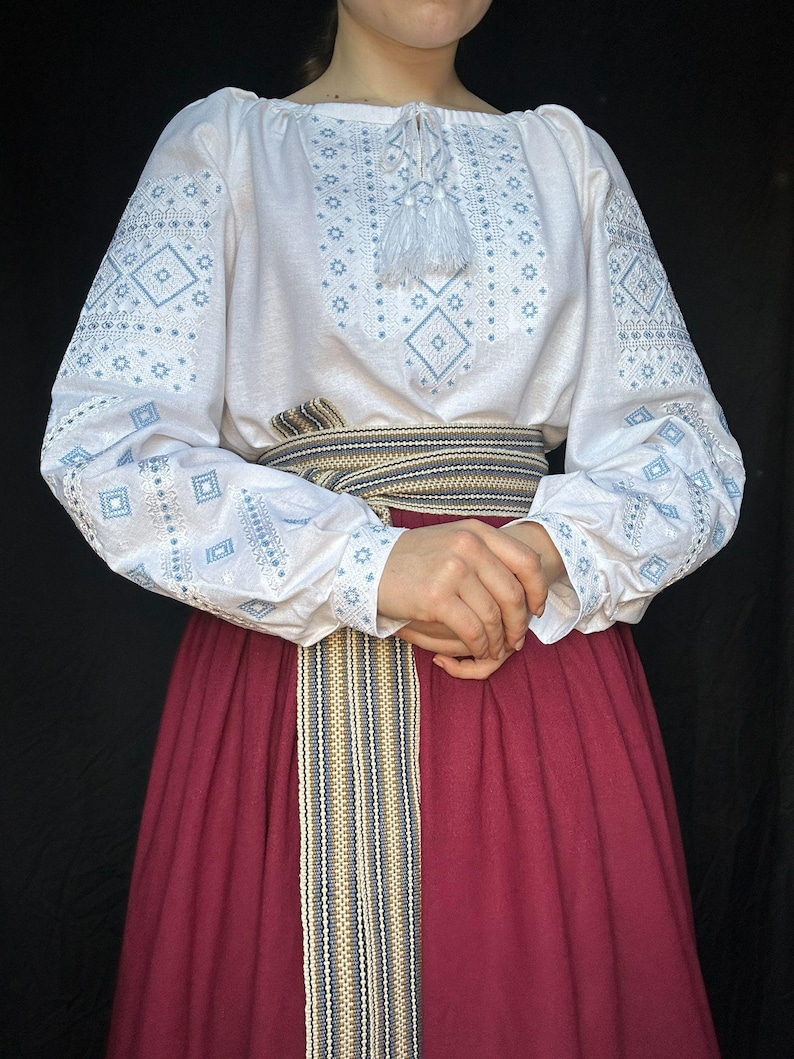 Puede incluir: Una blusa blanca bordada con motivos geom&eacute;tricos azules y una borla blanca. La blusa se lleva con una falda color burdeos y un cintur&oacute;n tejido con rayas beige, azules y negras.