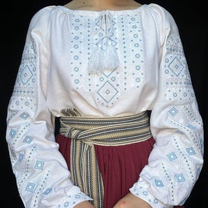 Puede incluir: Blusa blanca bordada con patrones geom&eacute;tricos azules y blancos, con un lazo de borla blanco en el cuello. La blusa se lleva con un cintur&oacute;n a rayas marr&oacute;n y blanco y una falda burdeos.