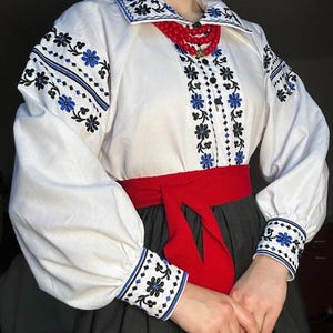 K&ouml;nnte beinhalten: Wei&szlig;e bestickte Bluse mit schwarzen und blauen Blumenmustern, einem roten G&uuml;rtel und einer roten Perlenkette.