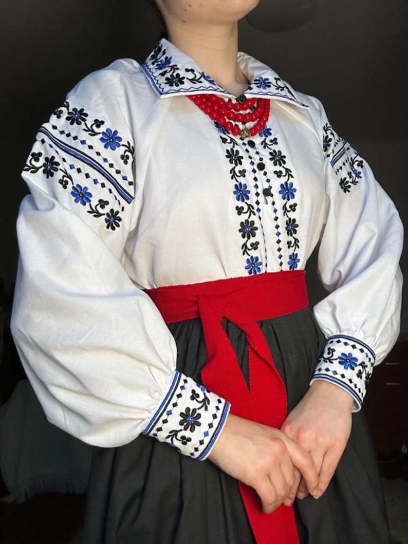 K&ouml;nnte beinhalten: Wei&szlig;e bestickte Bluse mit schwarzen und blauen Blumenmustern, einem roten G&uuml;rtel und einer roten Perlenkette.