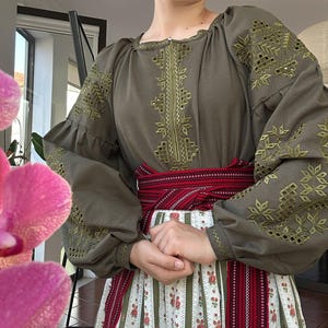 Può includere: Una camicetta a maniche lunghe verde oliva con intricati ricami geometrici color oliva sul petto e sulle maniche, abbinata a una gonna bianca con motivi floreali rossi e un'ampia fascia tessuta rossa e nera legata in vita.