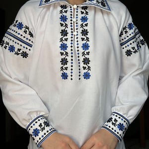 K&ouml;nnte beinhalten: Wei&szlig;es Langarmhemd mit blauen und schwarzen gestickten Blumenmustern an Kragen, Manschetten und Vorderseite.
