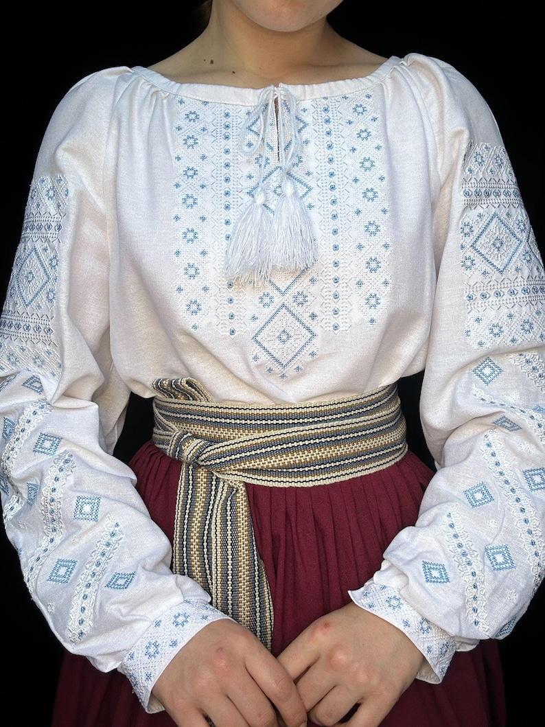 Puede incluir: Blusa blanca bordada con patrones geom&eacute;tricos azules y blancos. La blusa tiene una borla blanca en el escote y se lleva con un cintur&oacute;n a rayas marrones y blancas.