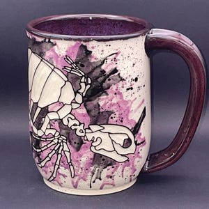 May include: A ceramic mug with a white base, featuring a black and white skeletal illustration and purple and black paint splatter design. The interior and handle are a deep purple colour. A unique and artistic coffee mug.