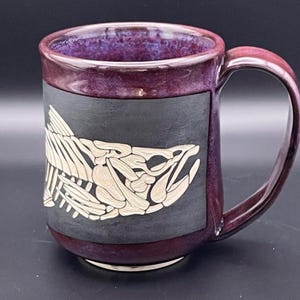 May include: A ceramic mug with a dark purple interior and handle. The exterior features a black rectangular panel with a cream-coloured fish skeleton design. The mug's base and rim are a deep burgundy colour.