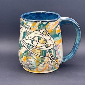 May include: A white ceramic mug with a teal blue handle and rim. The mug features a hand-painted design of a skeleton with a splatter paint background in shades of teal, yellow, and pink.