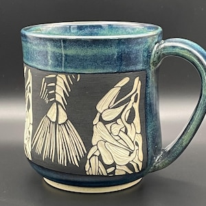 May include: Ceramic mug with a dark blue exterior and a teal interior. The mug features a black band with a cream-coloured fish skeleton design. The handle is teal and the mug is approximately 10 cm tall.