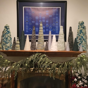 May include: A collection of decorative cone-shaped trees in various sizes and colors, arranged on a wooden shelf. The trees are made from textured materials in shades of green, white, gray, and brown. A garland with lights and greenery decorates the fireplace below.