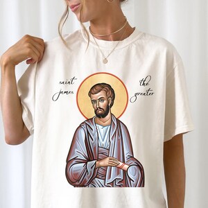 May include: A white t-shirt with a graphic of Saint James the Greater, a religious icon. The text "Saint James" and "The Greater" is printed on the shirt.