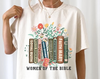 May include: A white t-shirt with a graphic of a stack of books with names of women from the Bible, such as Faith Mary, Patience Sarah, and Loyal Ruth. The graphic is surrounded by flowers. The text "Women of the Bible" is printed below the graphic.