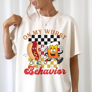 Può includere: Una t-shirt color bianco sporco con una stampa grafica di un hot dog e un boccale di birra in stile cartoon con il testo "ON MY WURST BEHAVIOR" su uno sfondo a scacchi bianco e nero. Il design include accenti arancioni, rossi e gialli.
