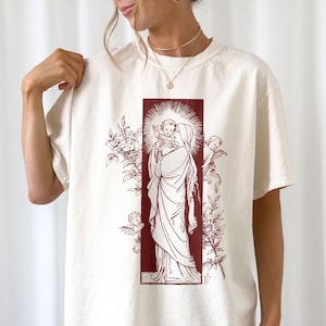 Può includere: T-shirt color crema con un'illustrazione bordeaux di una figura religiosa che tiene in braccio un bambino, incorniciata da un bordo rettangolare ed elementi floreali. Sono raffigurati anche due angeli. La maglietta ha maniche corte e una vestibilità ampia.