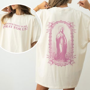 May include: A cream-colored oversized t-shirt with a pink graphic of the Virgin Mary on the back. The front of the shirt has the text "Mother Most Pure, Pray For Us!" in pink.