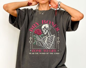 Pode incluir: Camiseta preta de tamanho grande com uma estampa gráfica de um esqueleto segurando uma rosa vermelha. O gráfico tem o texto "DRY BONES" em um círculo ao redor do esqueleto, com "EZ" e "37" de cada lado do círculo. O texto "COME TO LIFE" está abaixo do círculo, e o texto "HEAR THE WORD OF THE LORD" está abaixo disso.