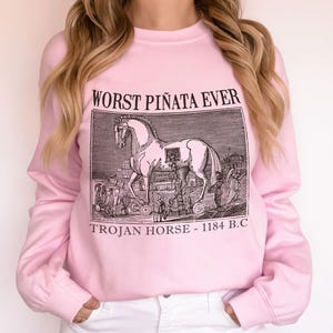 May include: Pink long-sleeve sweatshirt with the text "WORST PI&Ntilde;ATA EVER" above a black and white illustration of the Trojan Horse. The text "TROJAN HORSE - 1184 B.C" is below the illustration.