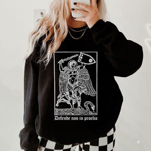 May include: Black sweatshirt featuring a white graphic of a figure with wings, a sword, and a shield. The text "Defende nos in proelio" is below the graphic. The sweatshirt has long sleeves and a crew neck.