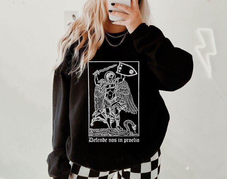 May include: Black sweatshirt featuring a white graphic of a figure with wings, a sword, and a shield. The text "Defende nos in proelio" is below the graphic. The sweatshirt has long sleeves and a crew neck.