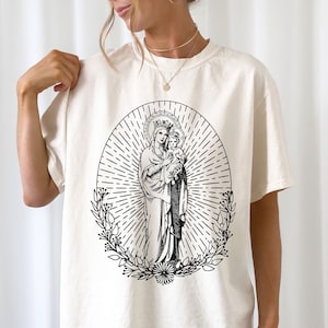 Può includere: Una maglietta bianca con un'illustrazione in bianco e nero della Vergine Maria e il bambino Gesù. L'illustrazione è circondata da una ghirlanda di fiori e un raggio di sole.