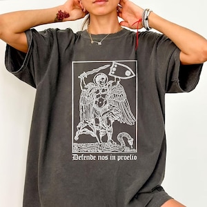 May include: Dark gray t-shirt featuring a white graphic of an angel with a sword and shield, and the text "Defende nos in proelio". The shirt has short sleeves and a relaxed fit.