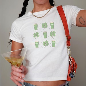 May include: White t-shirt featuring a pattern of green Guinness pints and four-leaf clovers. The design is repeated across the shirt. The shirt is short-sleeved and has a round neckline. The overall theme is St. Patrick's Day.