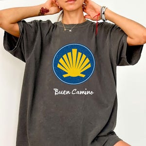 May include: A dark gray t-shirt with a blue and yellow scallop shell graphic and the text "Buen Camino" printed on the front.