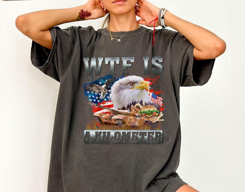 May include: Dark gray t-shirt with a graphic design featuring a bald eagle, American flag, and a tank holding a burger. The text "WTF IS A KILOMETER" is printed above and below the graphic.