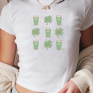 May include: White crop top featuring a repeating pattern of green Guinness beer glasses and four-leaf clovers. The design includes small green dots. The shirt is a casual, short-sleeved style.