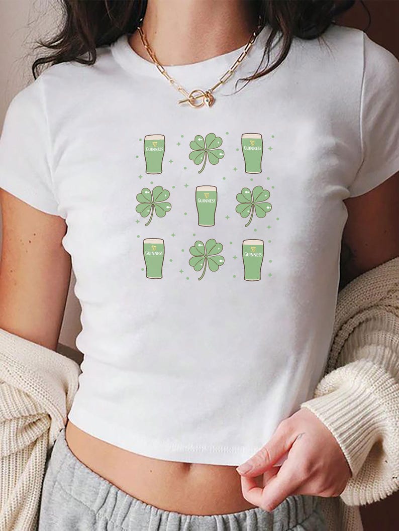 May include: White crop top featuring a repeating pattern of green Guinness beer glasses and four-leaf clovers. The design includes small green dots. The shirt is a casual, short-sleeved style.