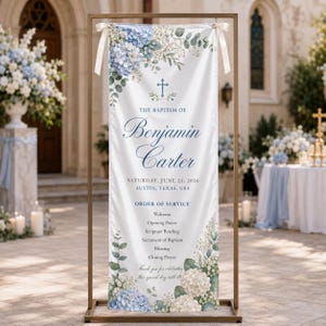 May include: A vertical white fabric banner suspended from a gold metal stand, featuring blue hydrangea and eucalyptus floral illustrations. The text reads: The Baptism of Benjamin Carter, Saturday, June 21, 2026, Austin, Texas, USA, Order of Service.