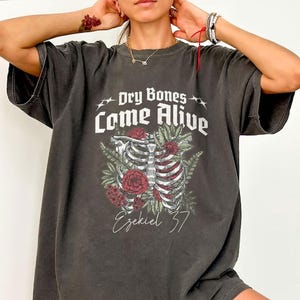 Peut inclure: Un t-shirt gris anthracite à manches courtes avec une impression graphique d'une cage thoracique humaine entourée de roses rouges et de feuilles vertes. Le texte "Dry Bones Come Alive" est imprimé en blanc au-dessus, avec "Ezekiel 37" en dessous.