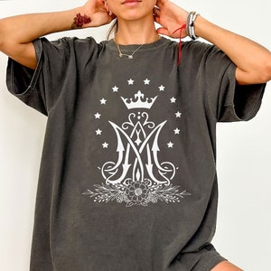 May include: An oversized t-shirt in a dark grey colour with a white graphic of a crown, a stylised "M" with a floral design, and a circle of stars.