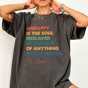 May include: Charcoal grey t-shirt featuring the quote "UNHAPPY IS THE SOUL ENSLAVED BY THE LOVE OF ANYTHING THAT IS MORTAL" in various colours. The quote is attributed to St Augustine. The shirt has a loose fit and short sleeves.