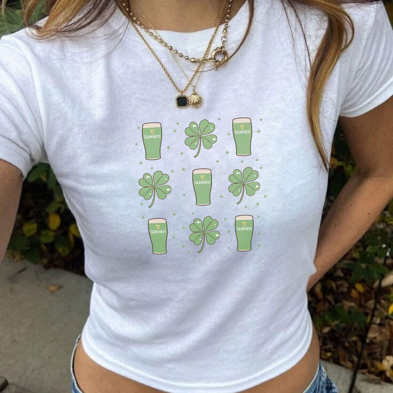 May include: White t-shirt featuring a pattern of green shamrocks and pints of Guinness. The design is arranged in a grid pattern. The shirt is a casual, everyday wear item, perfect for St. Patrick's Day celebrations.