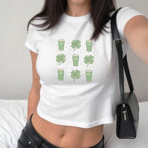 Può includere: Top corto bianco con un motivo ripetuto di bicchieri di birra verdi e quadrifogli. Il design è un'allusione giocosa al giorno di San Patrizio. La maglietta è un capo casual per tutti i giorni.