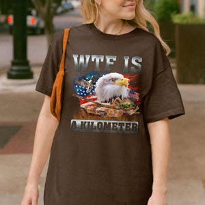 May include: Brown t-shirt featuring a graphic of an American bald eagle, the US flag, and a military tank. The text "WTF IS A KILOMETER" is printed across the front in a silver, distressed font.