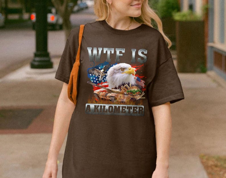 May include: Brown t-shirt featuring a graphic of an American bald eagle, the US flag, and a military tank. The text "WTF IS A KILOMETER" is printed across the front in a silver, distressed font.