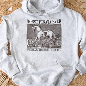 May include: Light gray hoodie with a front pocket and drawstring hood. The graphic on the front features a black and white illustration of the Trojan Horse with the text "WORST PI&Ntilde;ATA EVER" and "TROJAN HORSE - 1184 B.C."