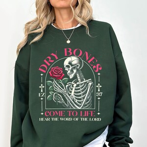 May include: Dark green sweatshirt with a graphic print of a skeleton holding a rose. The text "DRY BONES" is written in pink above the skeleton and "COME TO LIFE" is written below the skeleton. The text "HEAR THE WORD OF THE LORD" is written below the graphic.