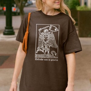 May include: A dark brown t-shirt featuring a white graphic of an angel with a sword and shield, and the text "Defende nos in proelio" in a rectangular frame. The shirt has short sleeves and a relaxed fit.